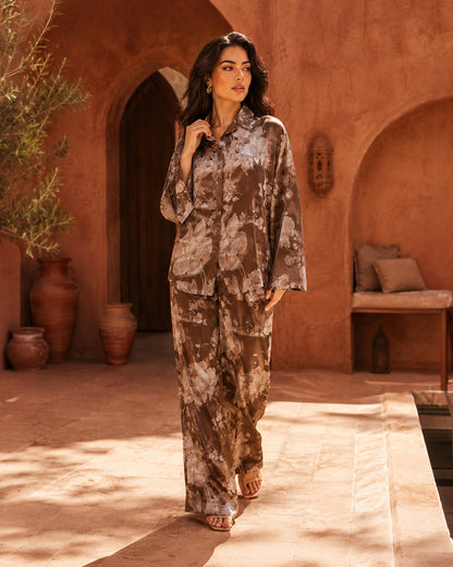 Model walking in a courtyard wearing the Sahara Bloom bronze floral silk shirt and matching wide-leg trousers.